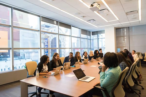 A diverse group of real estate professionals gathered around a large wooden conference table in a bright, modern office with floor-to-ceiling windows, collaborating on laptops.