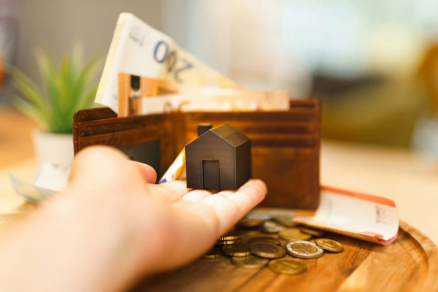 Hand holding a miniature house in front of a wallet with Euro bills and coins, symbolizing home valuation, property investment, and real estate financing. How Much Is Your House Worth?